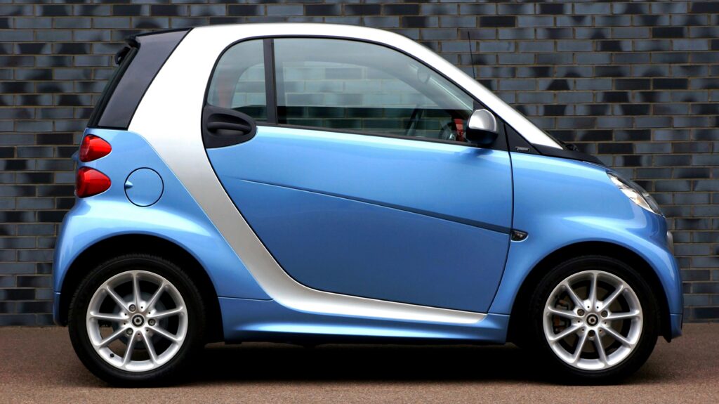 Side view of a modern blue smart car against a brick wall, showcasing compact design.